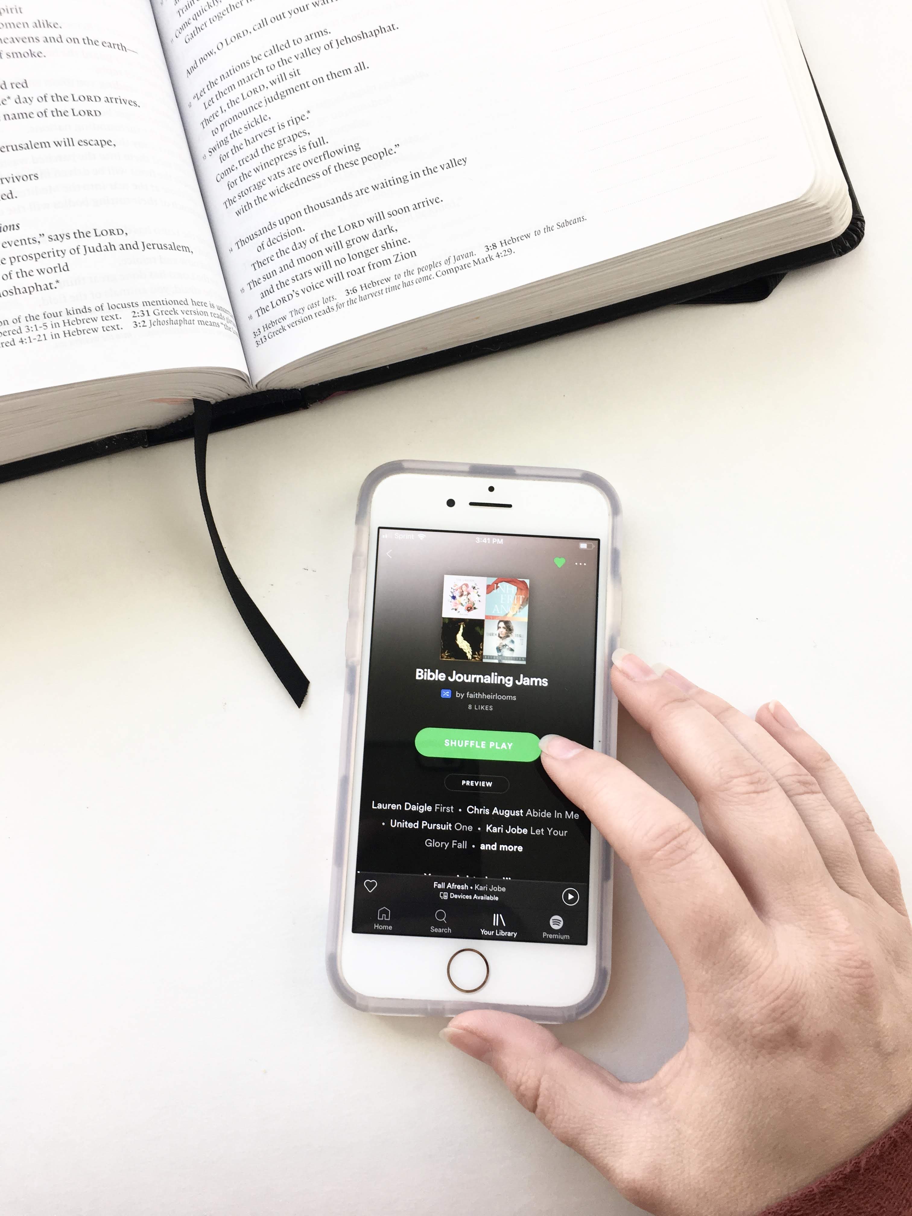 Bible and iPhone. Girl's hand clicking Bible Journaling Jams playlist on Spotify.